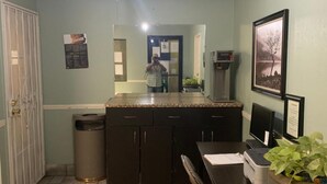 A compact kitchen with a tiled floor, a ceiling fan, a trash bin, and a dining area with a table and chairs. - Palms Inn & Suites (Palmdale)