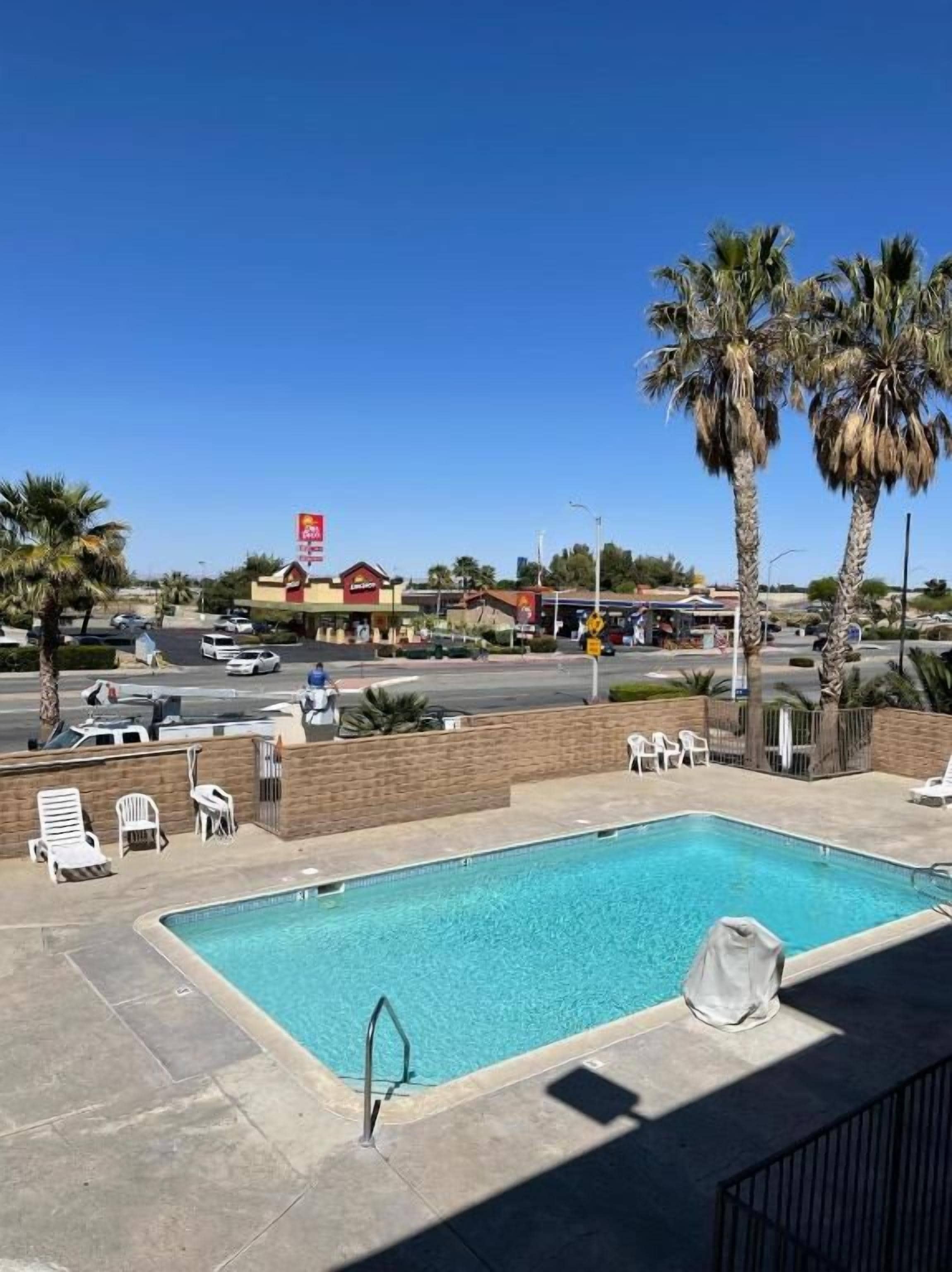 A swimming pool with lounge chairs, palm trees, and a view of a commercial area with a red sign.