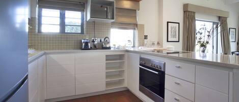 Een moderne keuken met witte kastjes, een inbouwoven en een magnetron. Er is een raam met gordijnen te zien, en buiten staat een plant.