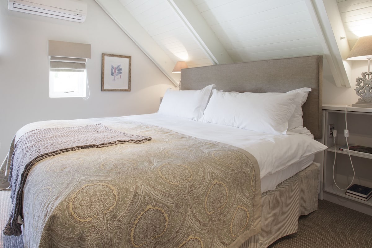 Un dormitorio con una cama grande, un cabecero, una mesita de noche con lámpara y una ventana con persianas.