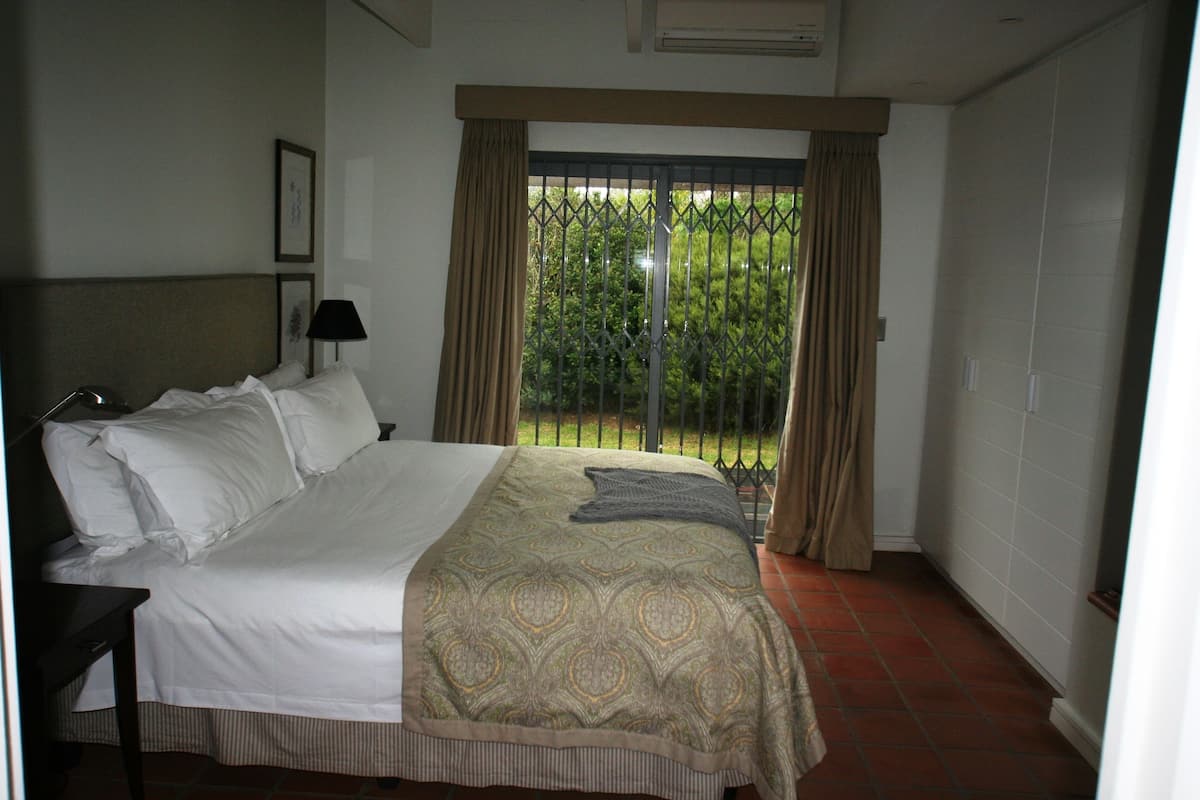 Un cuarto de hotel con una cama grande, una mesita de noche, una lámpara y vistas de un jardín a través de una ventana con barrotes.