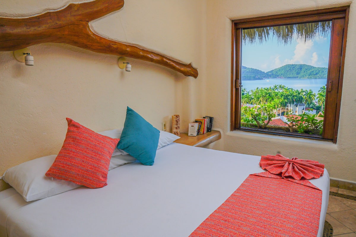 Una cama con una colcha blanca y roja, dos almohadas, y vista a un lago y palmeras a través de una ventana.