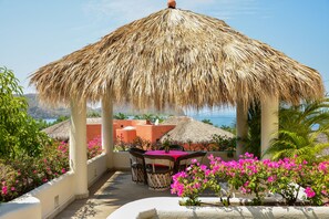 A covered outdoor seating area with a thatched roof, surrounded by vibrant pink flowers and palm trees, overlooking a scenic coastal view. - Villas San Sebastián (Zihuatanejo)
