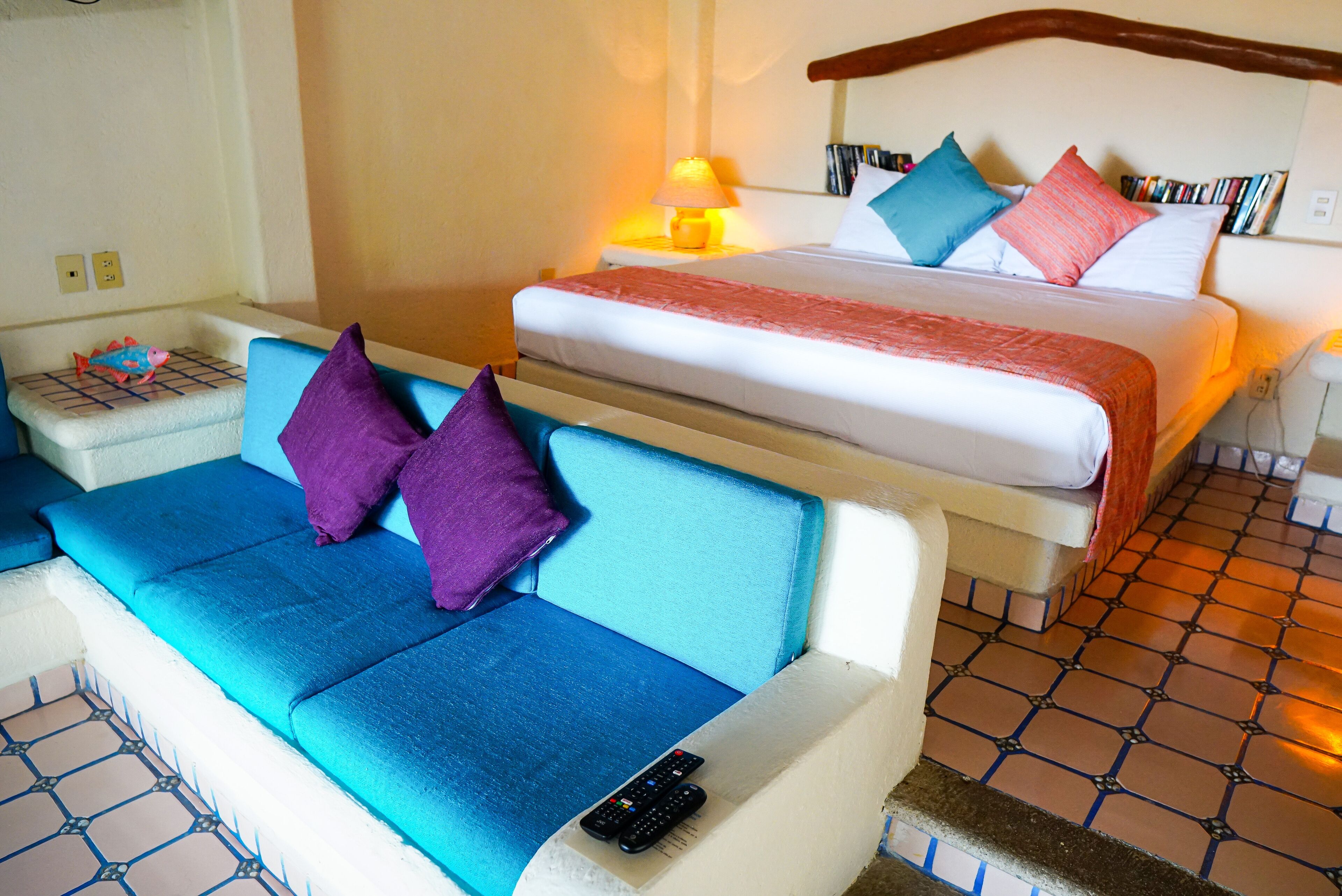 Habitación de hotel con cama, sofá azul, una mesita con lámpara y suelo de baldosas.