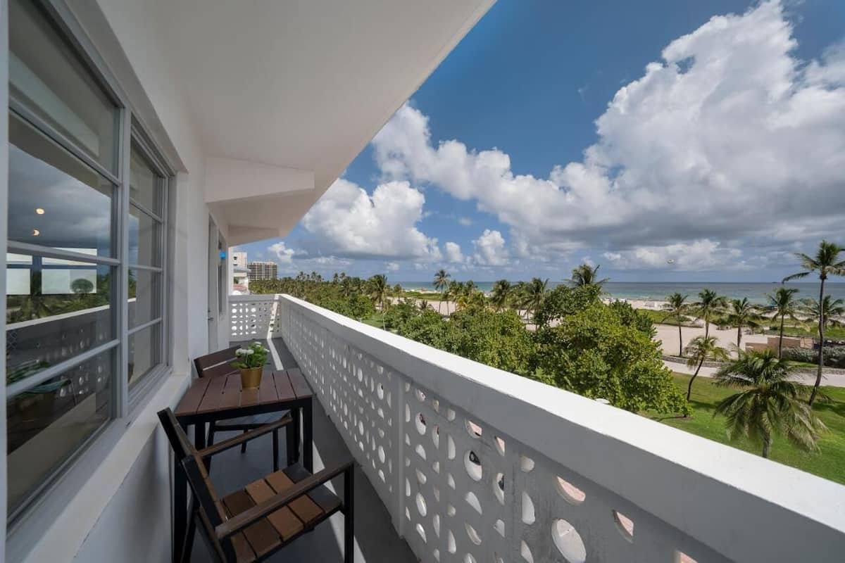 Un balcon avec une table et des chaises, donnant sur une plage bordée de palmiers et sous un ciel dégagé.