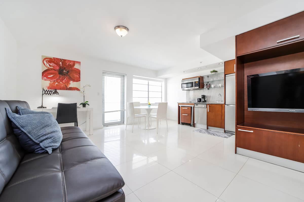 Un salon moderne avec un canapé en cuir foncé, un téléviseur à écran plat et un coin repas comprenant une table et des chaises blanches.