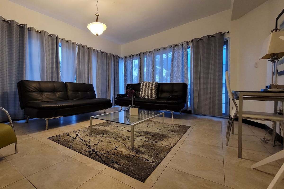 Un salon avec deux canapés en cuir noir, une table basse en verre, un tapis à motifs et un coin repas comprenant une table et des chaises.