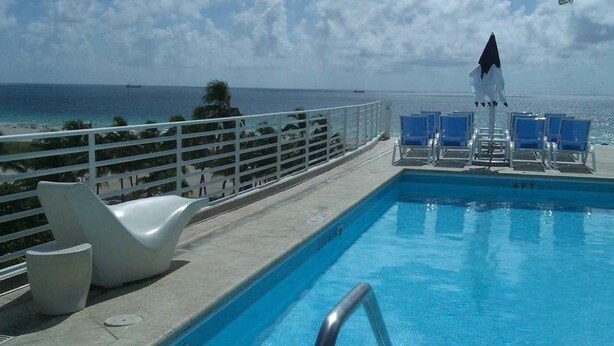 A pool with a view of the ocean, lounge chairs, and a white umbrella. - Strand Ocean Drive Suites (Miami Beach)