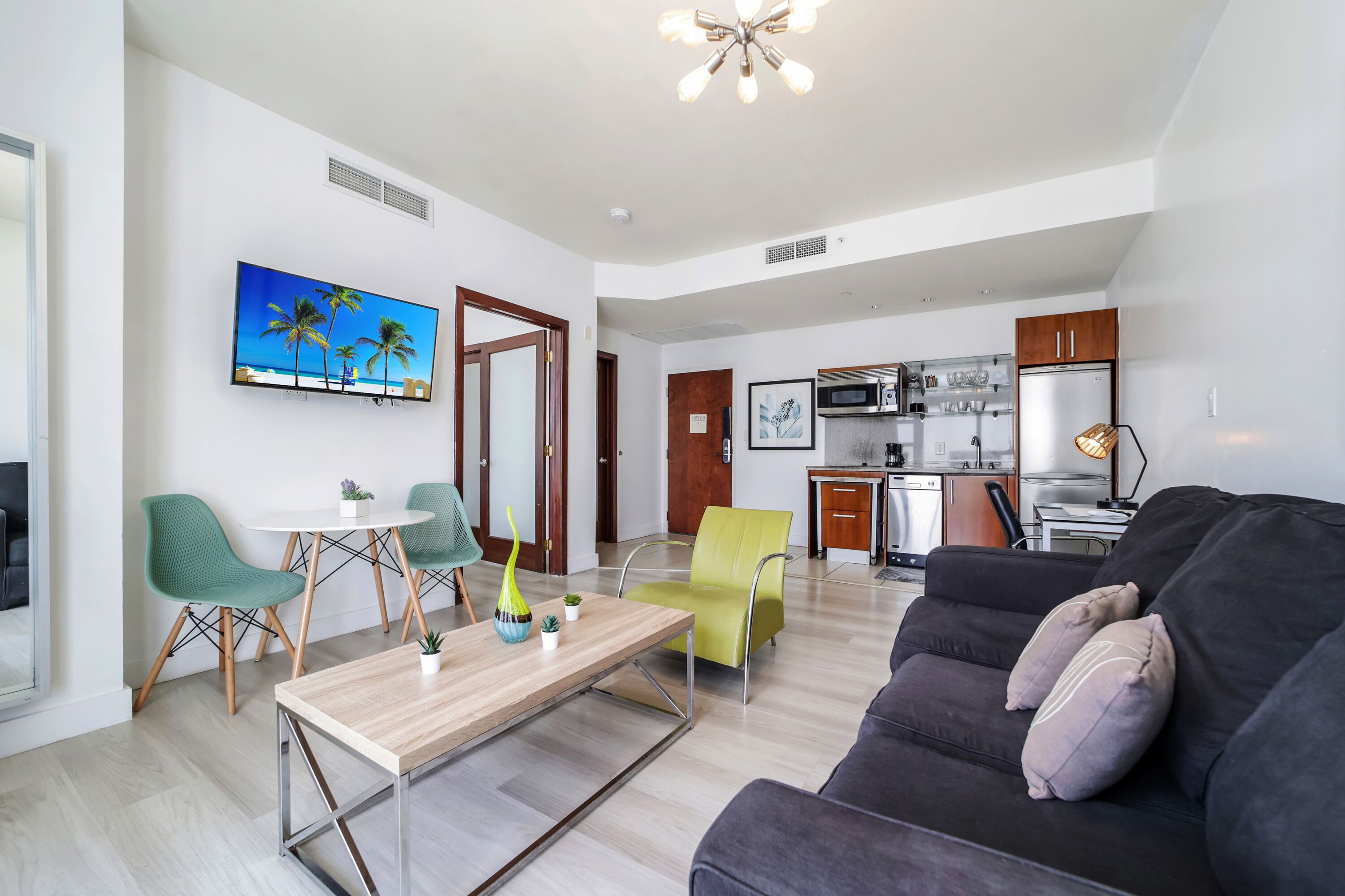 A modern living room with a sofa, chairs, a coffee table, and a TV. There is a kitchen area with a microwave and cabinets.