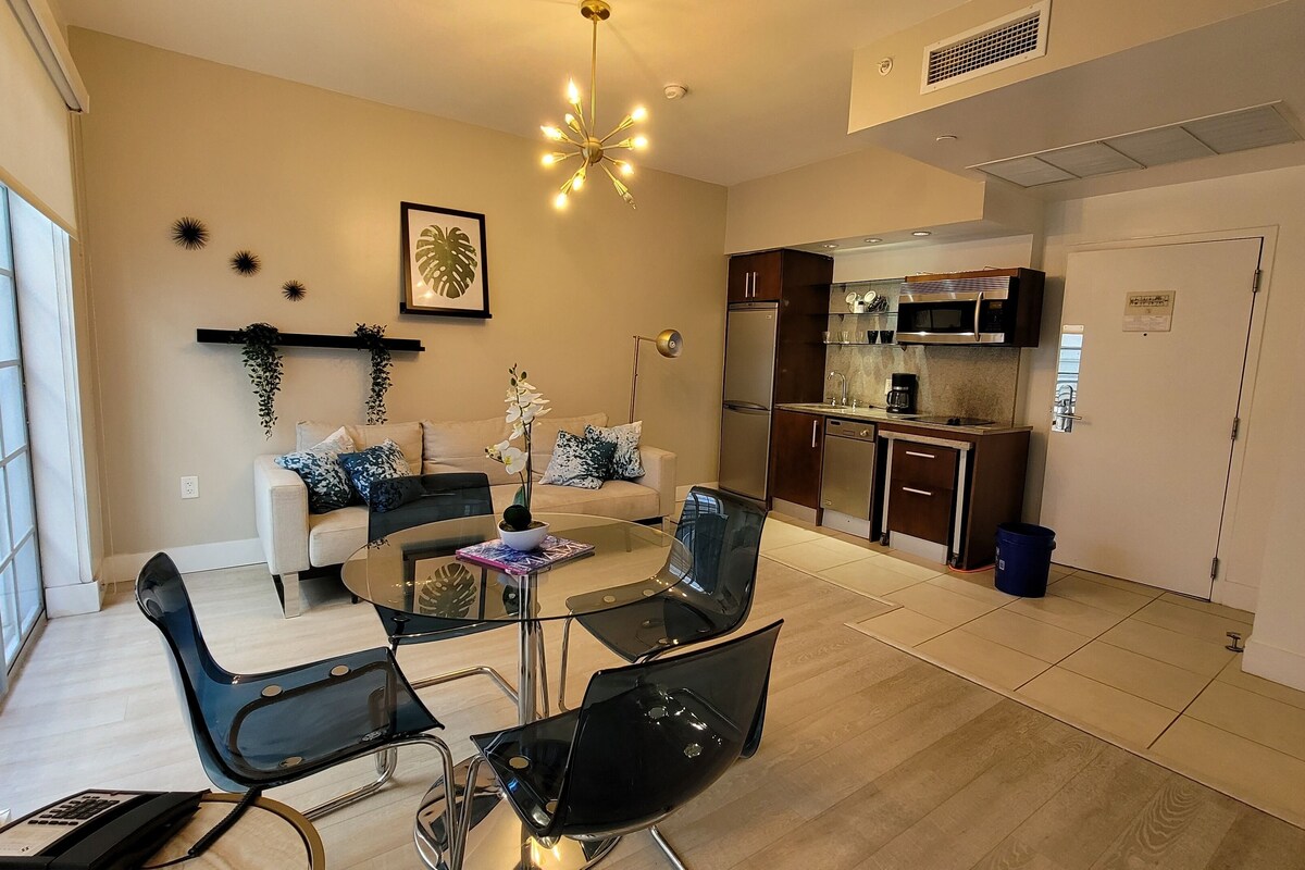 Una sala de estar moderna con una mesa de comedor de vidrio, sillas negras, un sofá y una zona de cocina con microondas y refrigerador.