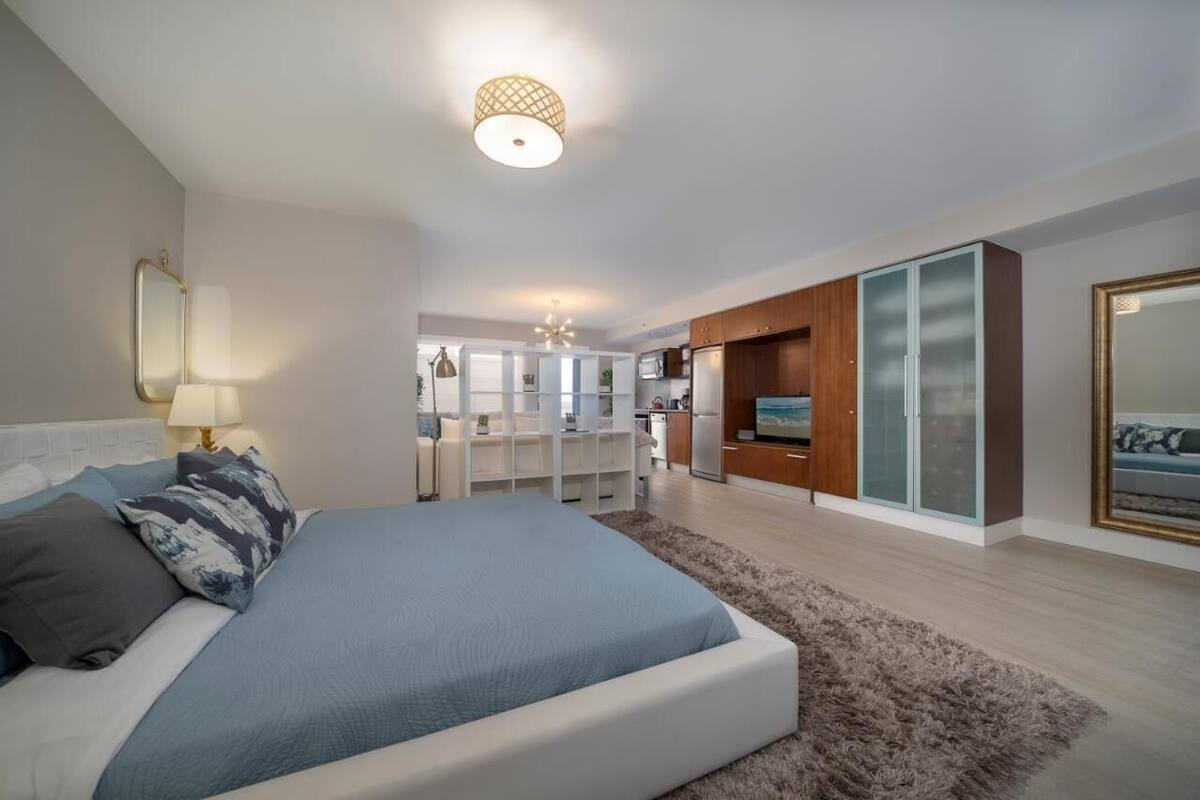 Une chambre moderne avec un grand lit, une télévision et un coin cuisine à l’arrière-plan.