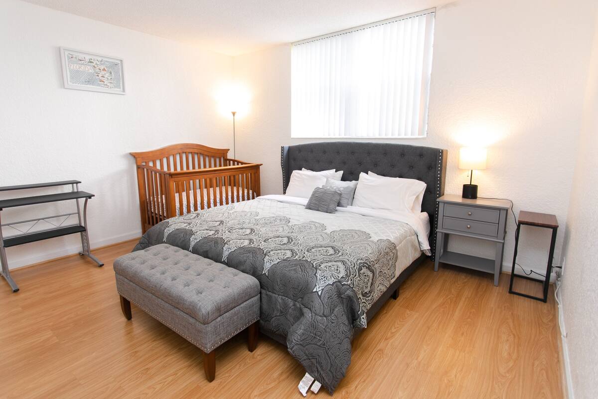 A bedroom with a large bed, a wooden crib, a grey upholstered bench, a nightstand, and a small table.
