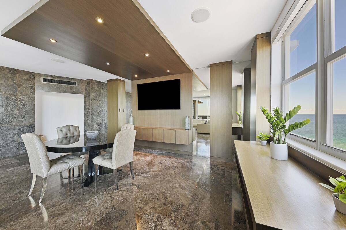 A modern living room with a large TV, a dining area with a round table and chairs, and a view of the ocean through large windows.