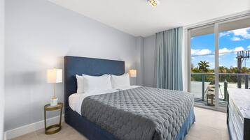 A modern hotel room with a large bed, a nightstand, a lamp, and a balcony with a view of the sky and palm trees.