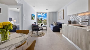 A modern kitchen with a marble countertop, a dining area with a round table and chairs, a sofa, and a view through a glass door to the outdoors.