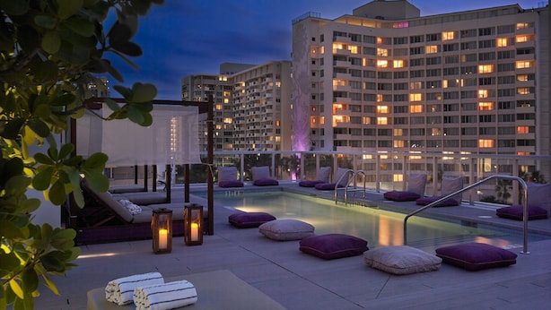 A rooftop pool area with lounge chairs and a jacuzzi, surrounded by a high-rise building with illuminated windows.