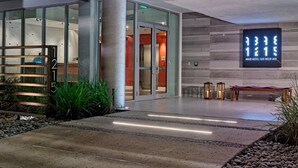 A modern hotel entrance with a glass facade, a wooden bench, and a sign indicating the hotel's name and address.