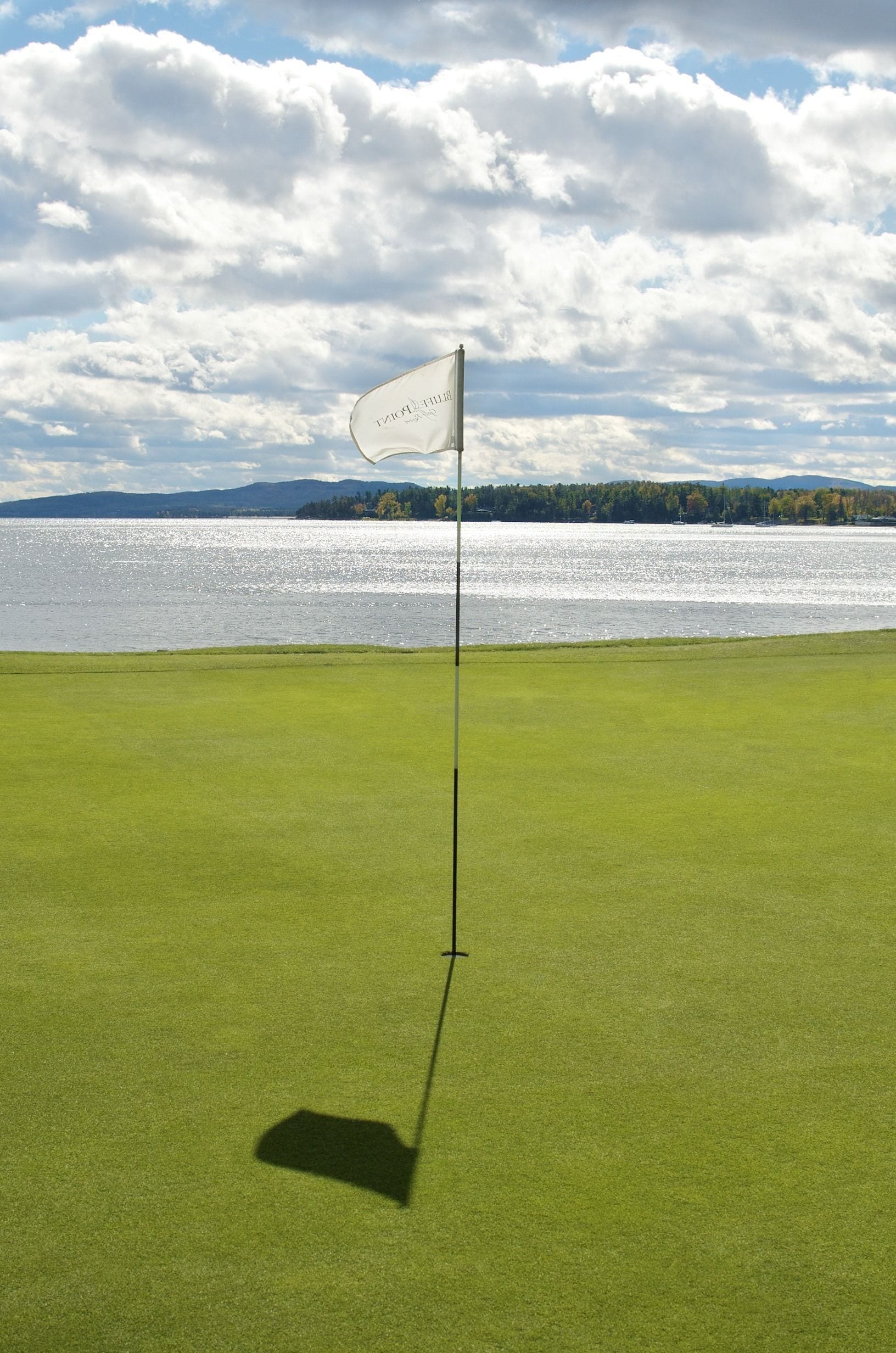 A golf course with a flagstick on a green, a body of water, and a cloudy sky in the background.