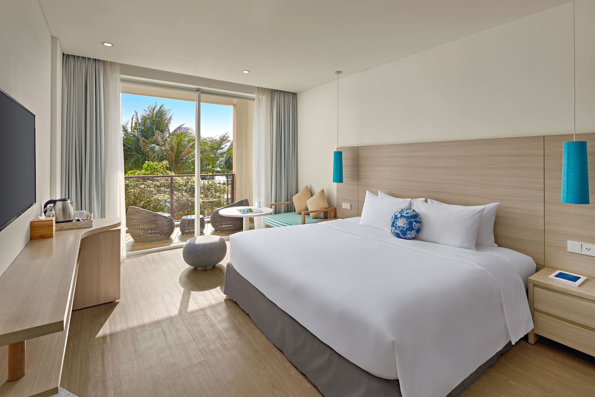 A modern hotel room with a large bed, a desk, a chair, and a balcony with a view of palm trees.