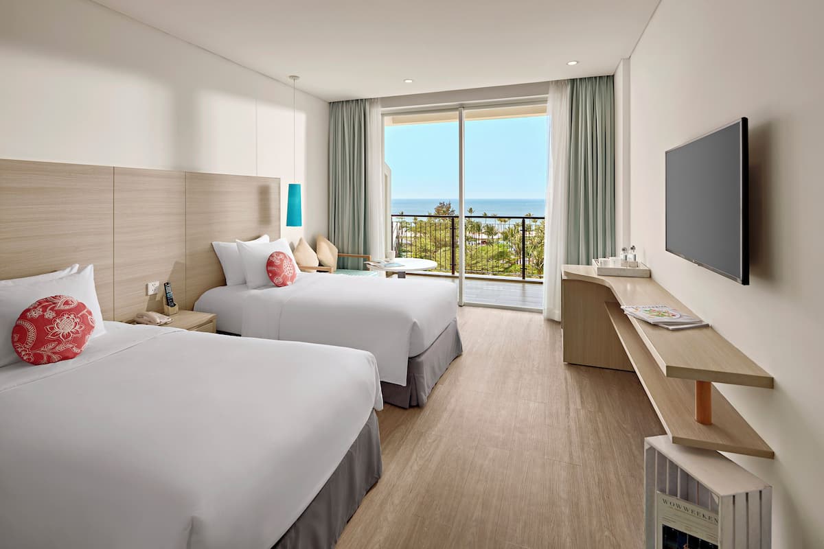 A hotel room with two beds, a desk, a TV, and a balcony with a view of the ocean.