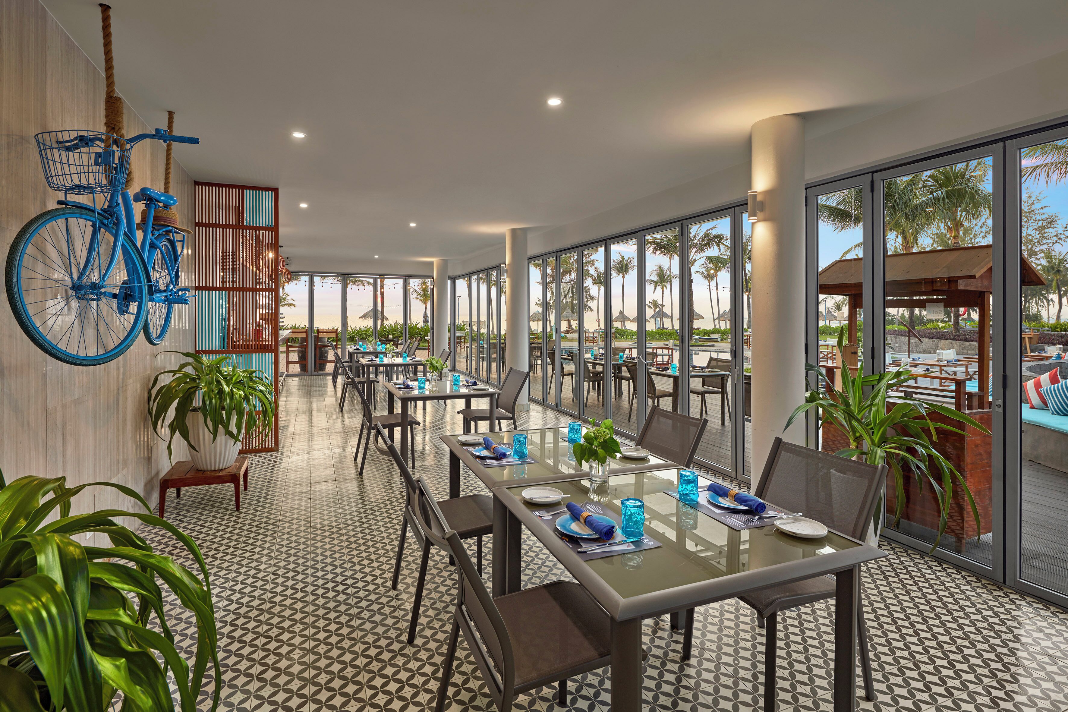 A spacious dining area with large windows, a bicycle mounted on the wall, and several tables set for dining.