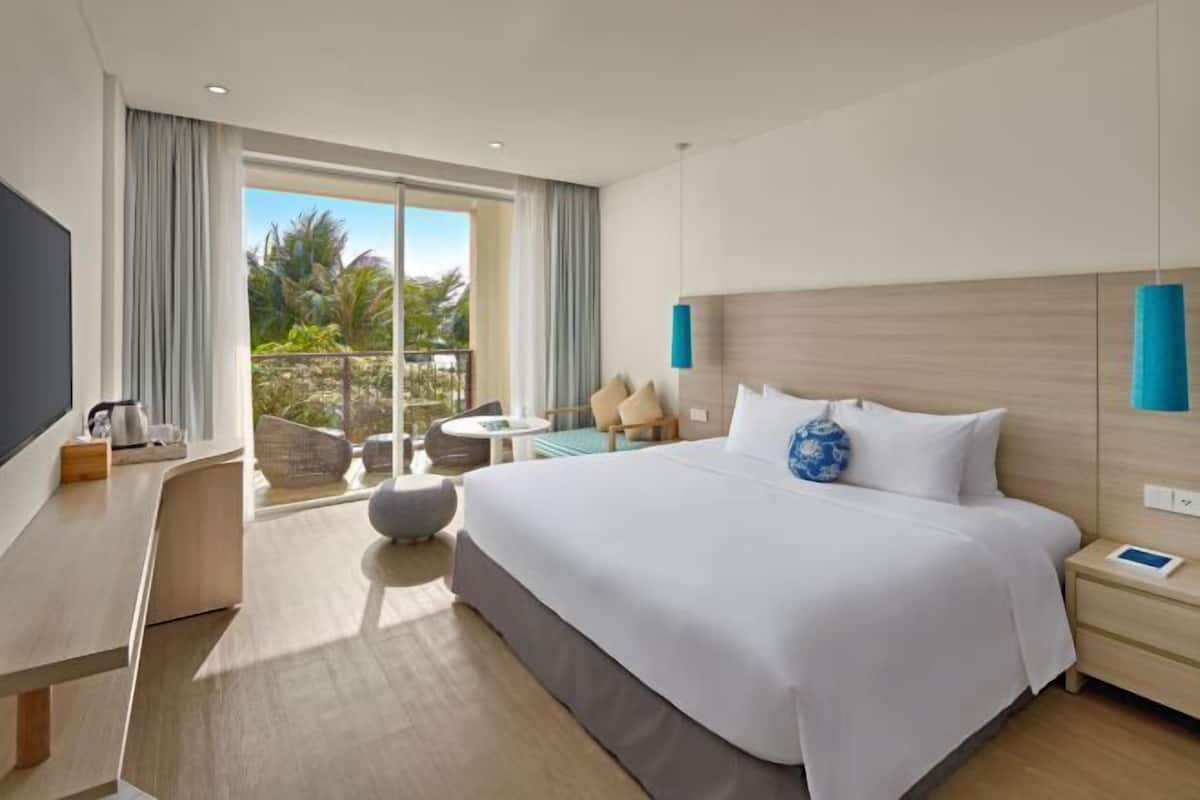 A modern hotel room with a large bed, a desk, a TV, and a balcony with a view of palm trees.