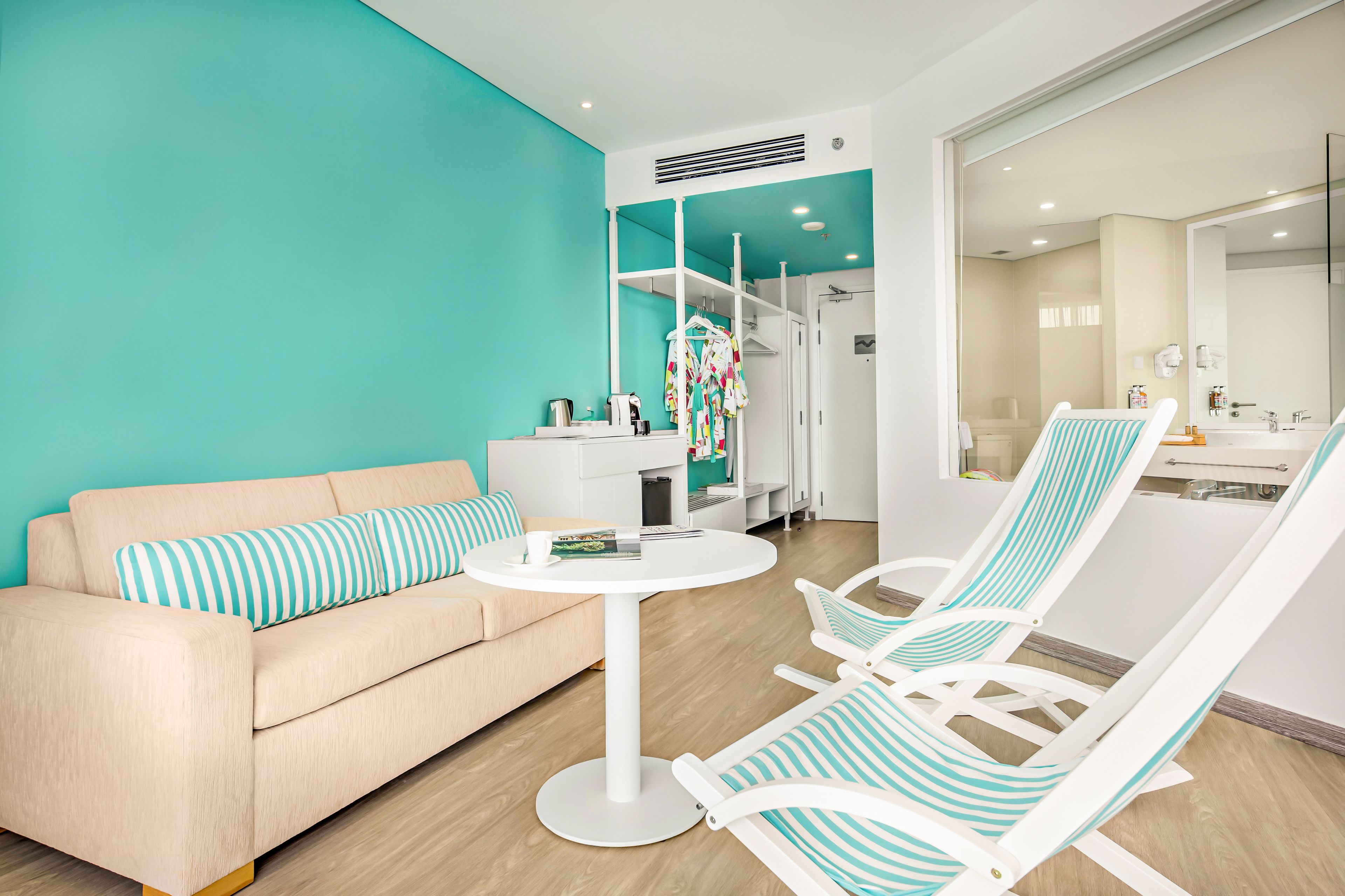 A modern living room with a teal accent wall, a beige sofa, a round white table, and two striped lounge chairs.