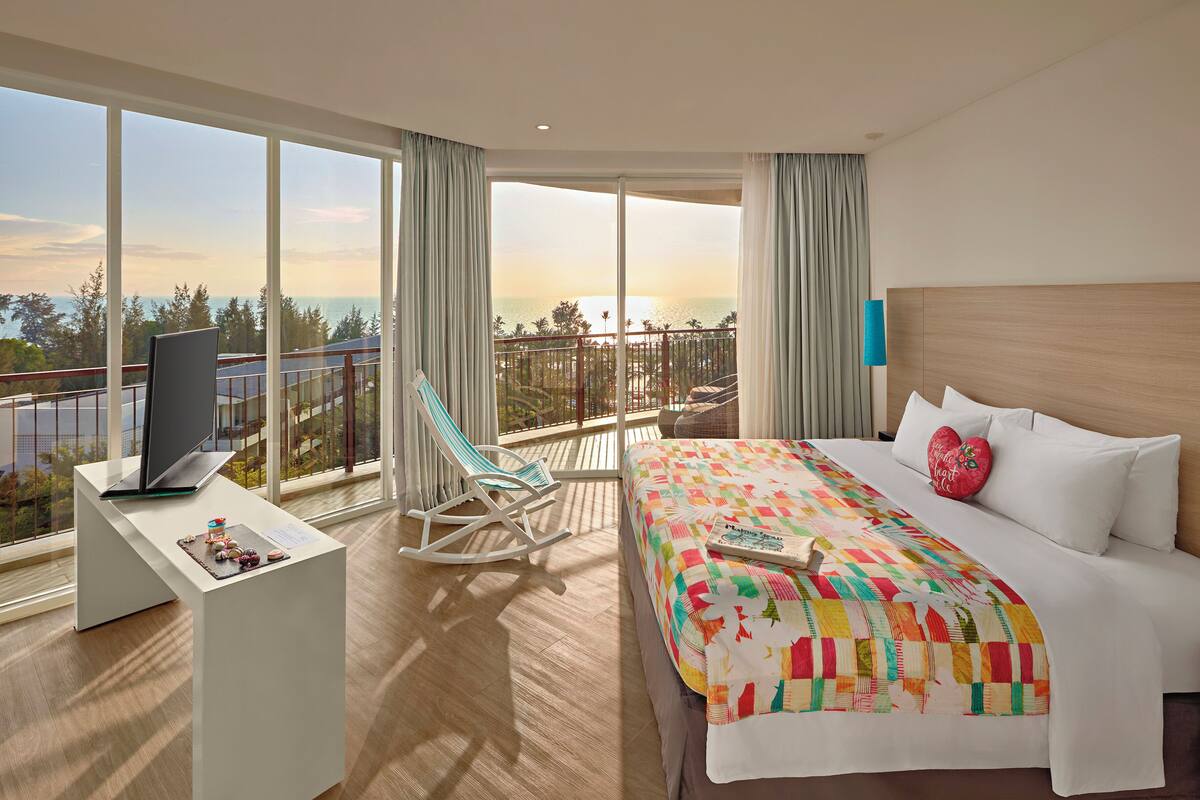 A hotel room with a large bed, a desk with a laptop, a rocking chair, and a balcony with a view of the ocean.