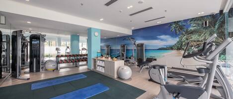 A modern gym with exercise equipment, blue mats, and a scenic mural of a beach.
