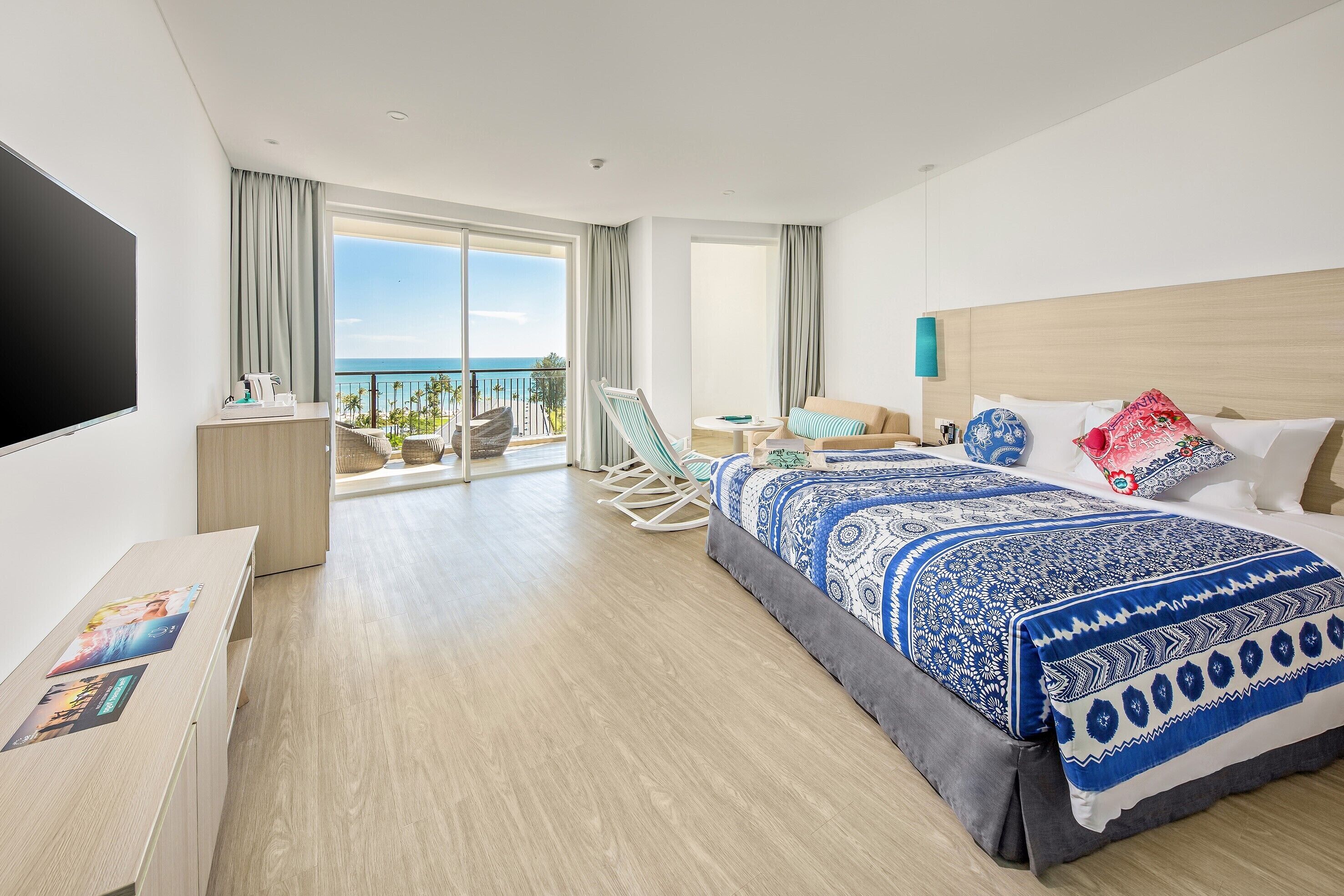 A modern hotel room with a large bed, a TV, a desk, and a balcony with a view of the ocean.