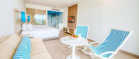 A modern hotel room with a bed, a sofa, a dining table with fruit, and a TV.