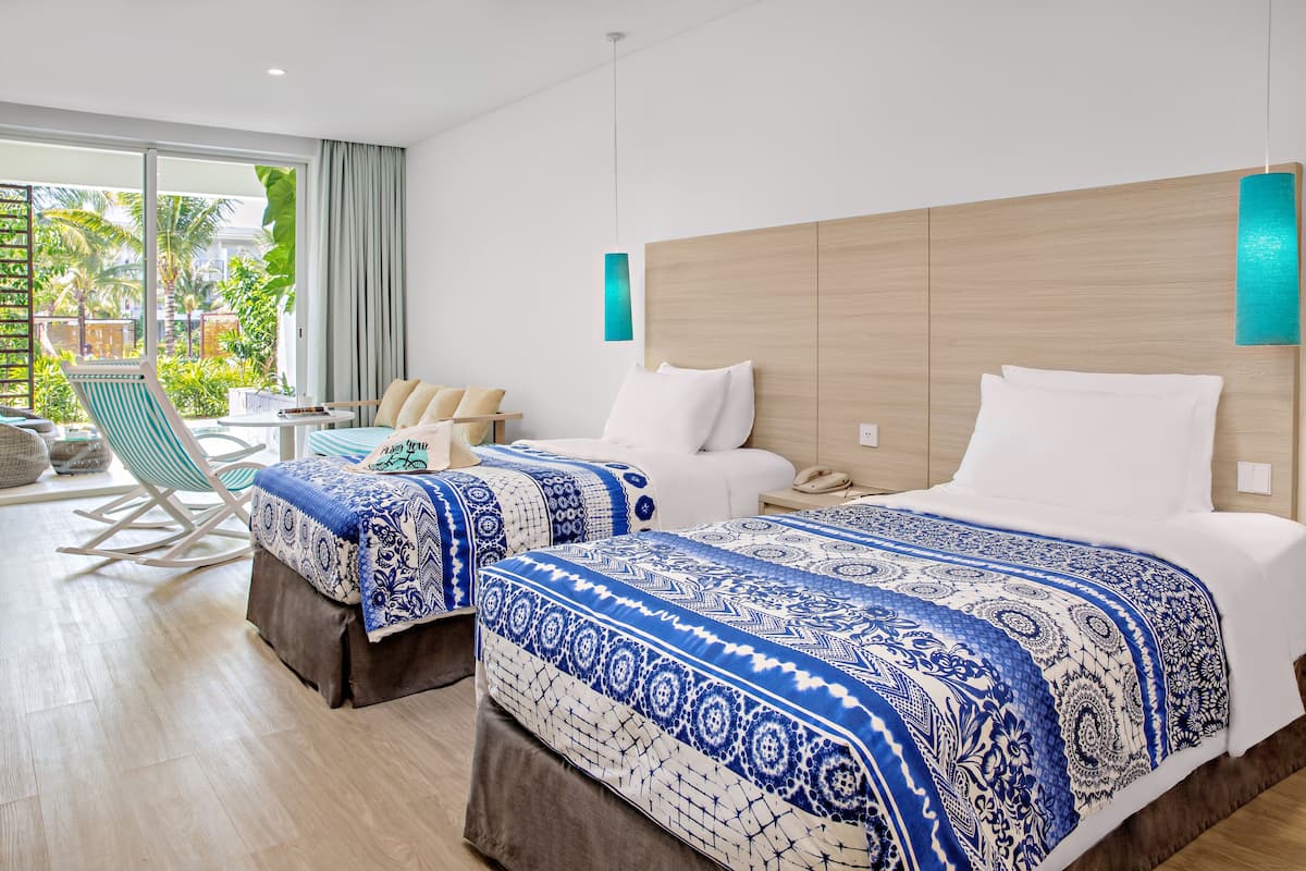 A hotel room with two beds, a sitting area with a chair, a small table, and a view of the outdoors.