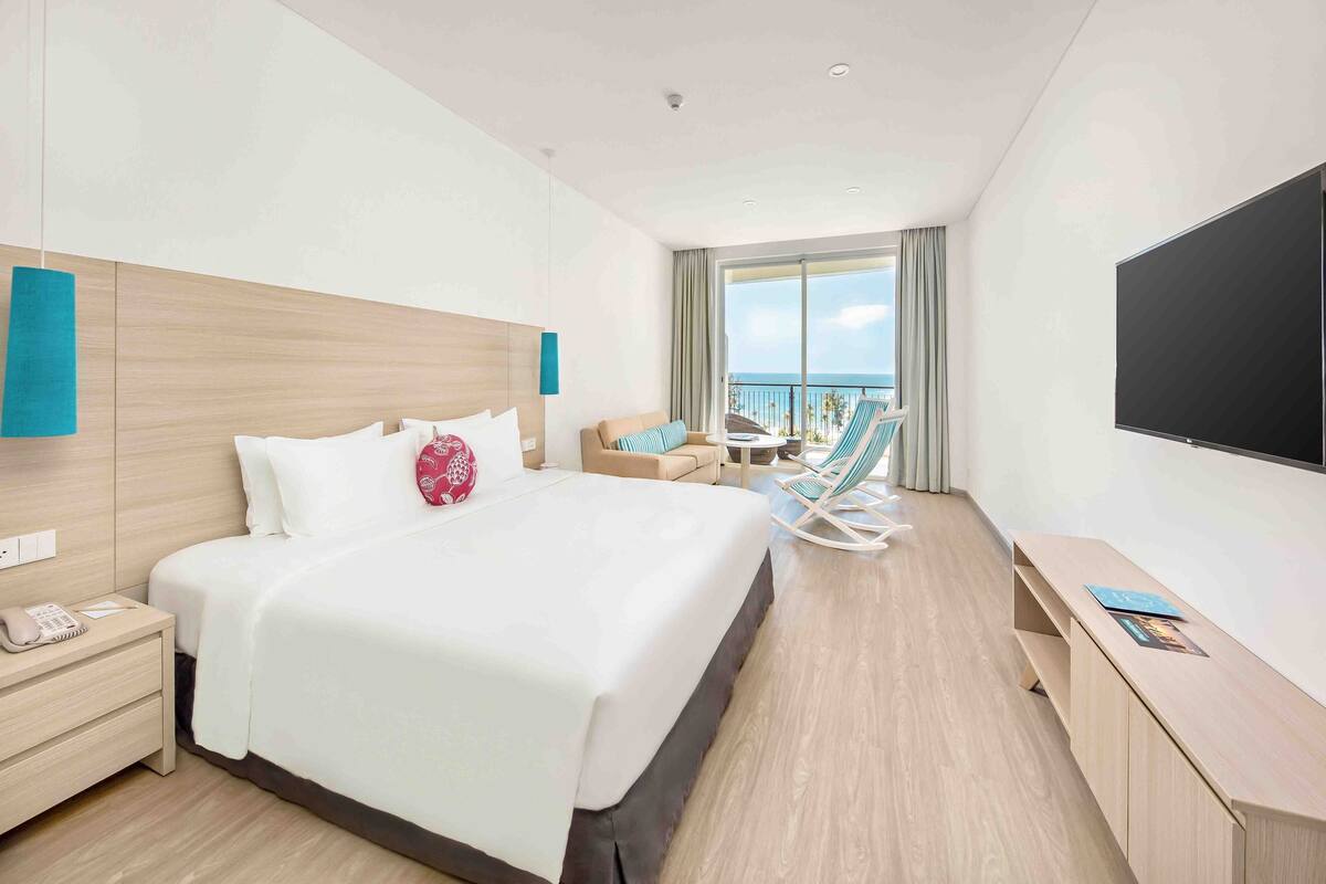 A hotel room with a large bed, a desk, a TV, and a balcony with a view of the sea.