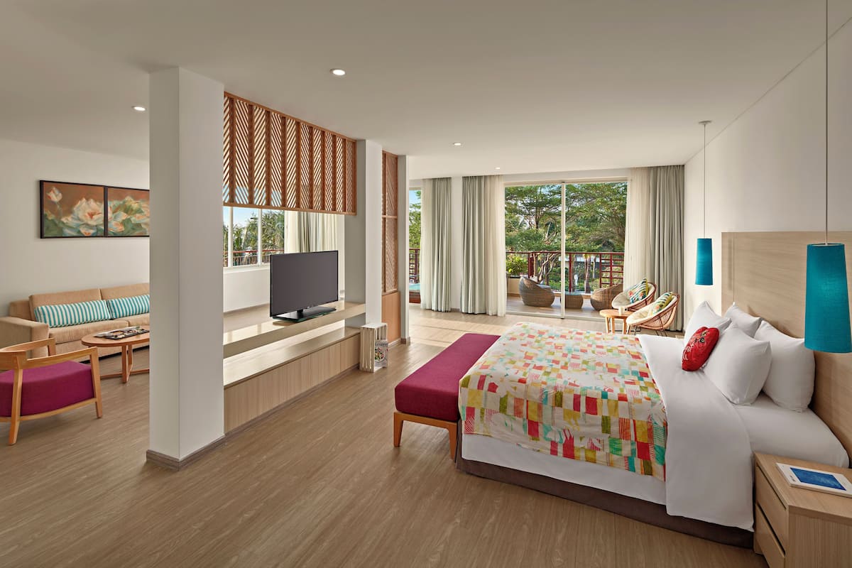 A modern hotel room with a large bed, a TV, a balcony with outdoor seating, and a view of greenery.