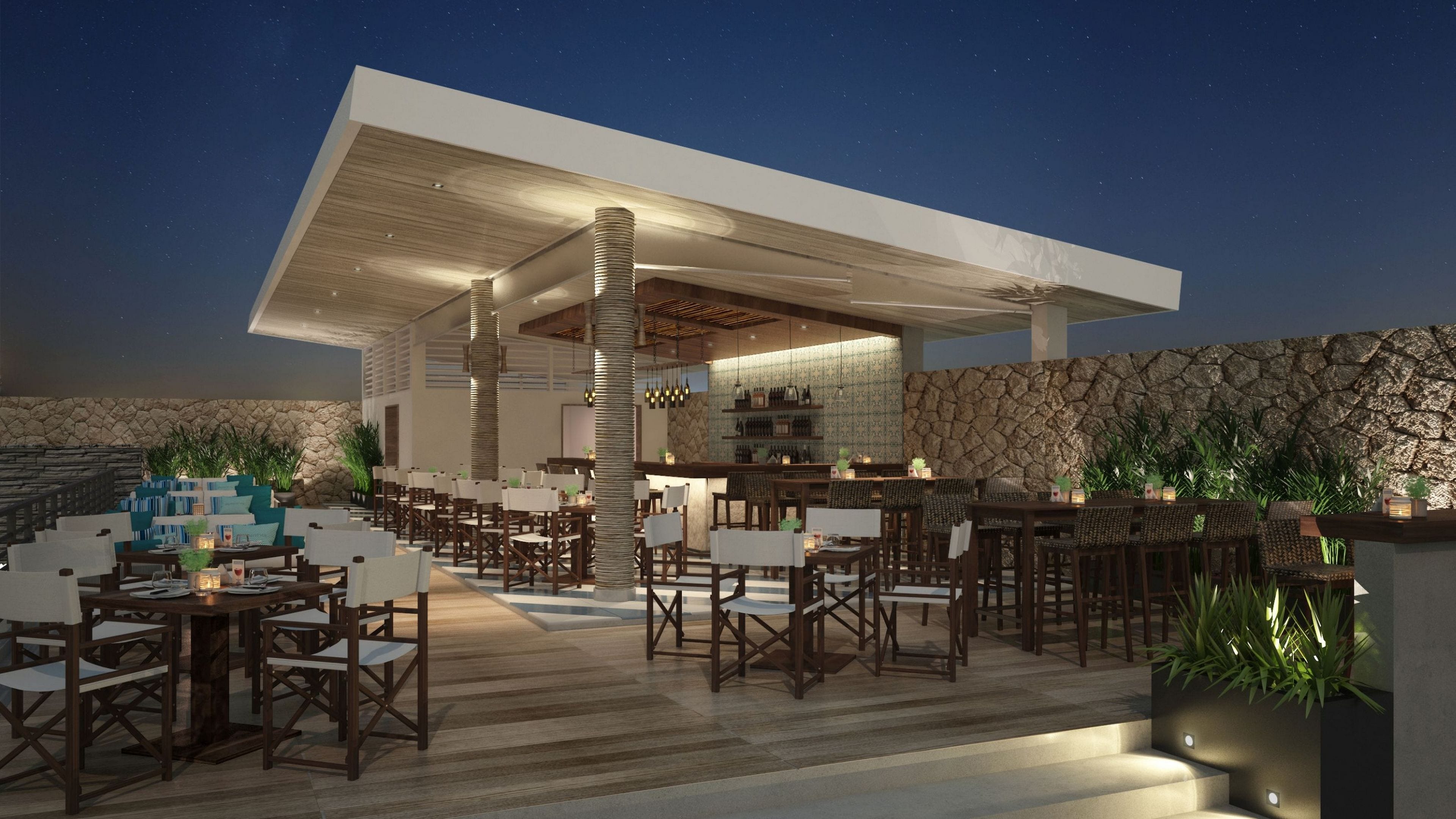 An outdoor dining area with multiple tables and chairs, a bar, and a stone wall backdrop.