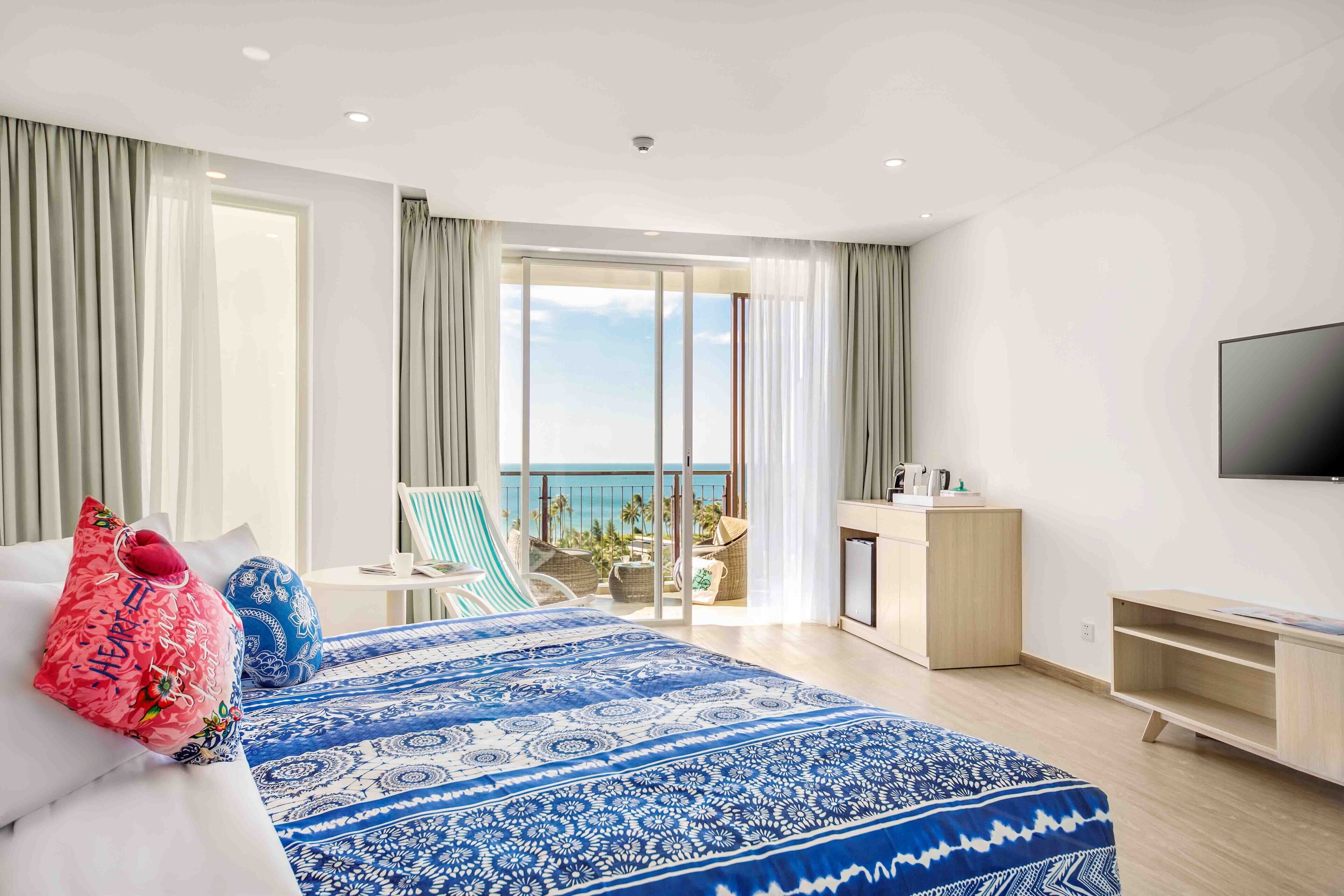 Ein Hotelzimmer mit einem großen Bett, einem Schreibtisch, einem Fernseher und einem Balkon mit Meerblick.