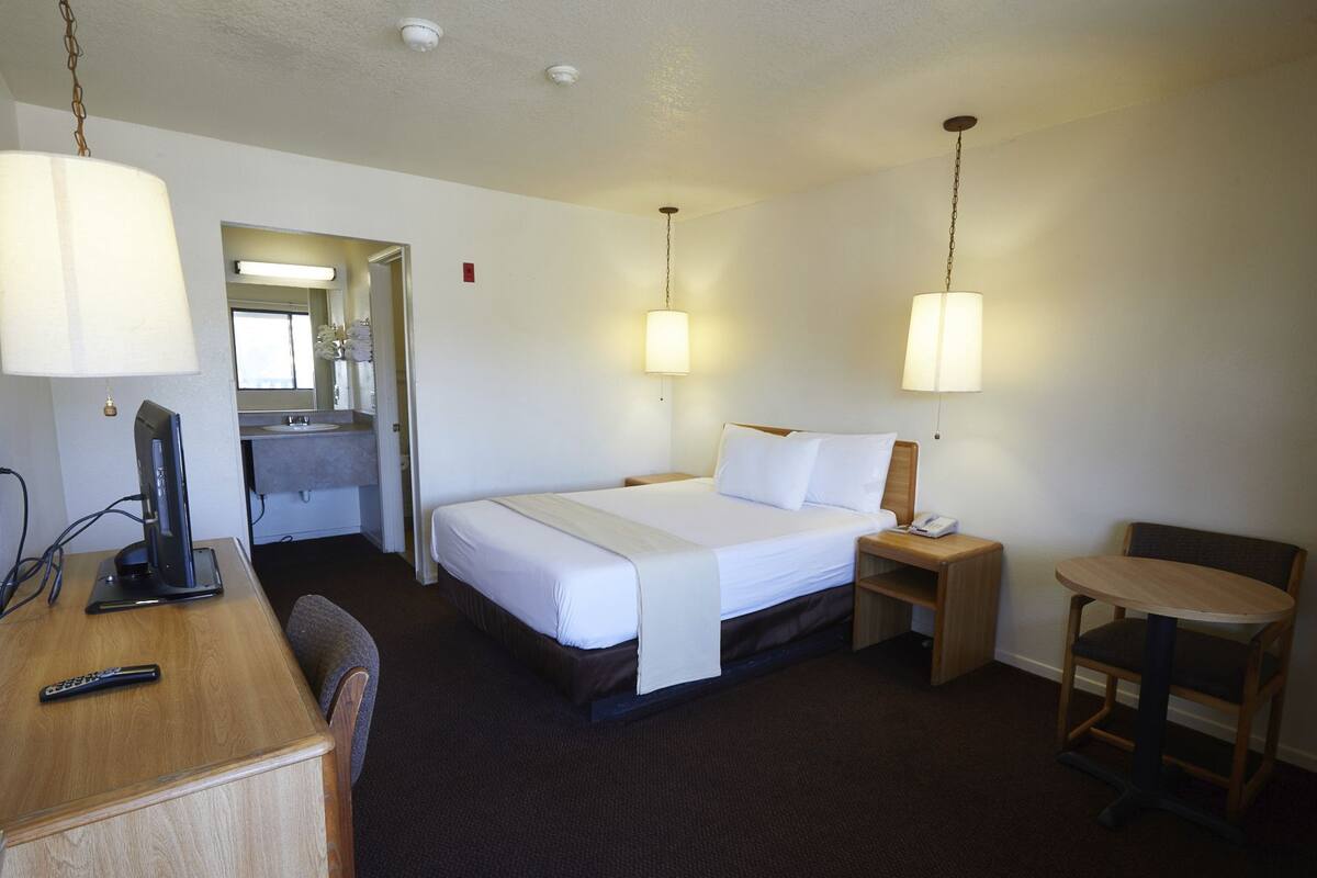 Ett hotellrum med en säng, ett sängbord, en stol, ett skrivbord med en dator och ett badrum med en spegel och ett handfat.