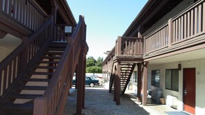 A building with a wooden staircase leading to a balcony, a parking area with cars, and a building entrance with a door marked "112".