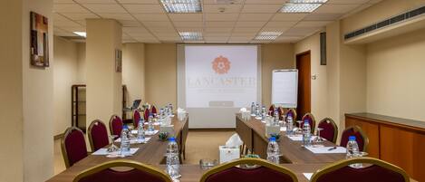 Un salón de conferencias con una larga mesa, sillas rojas, botellas de agua y una pantalla proyectora mostrando el logo del Lancaster Hotel.