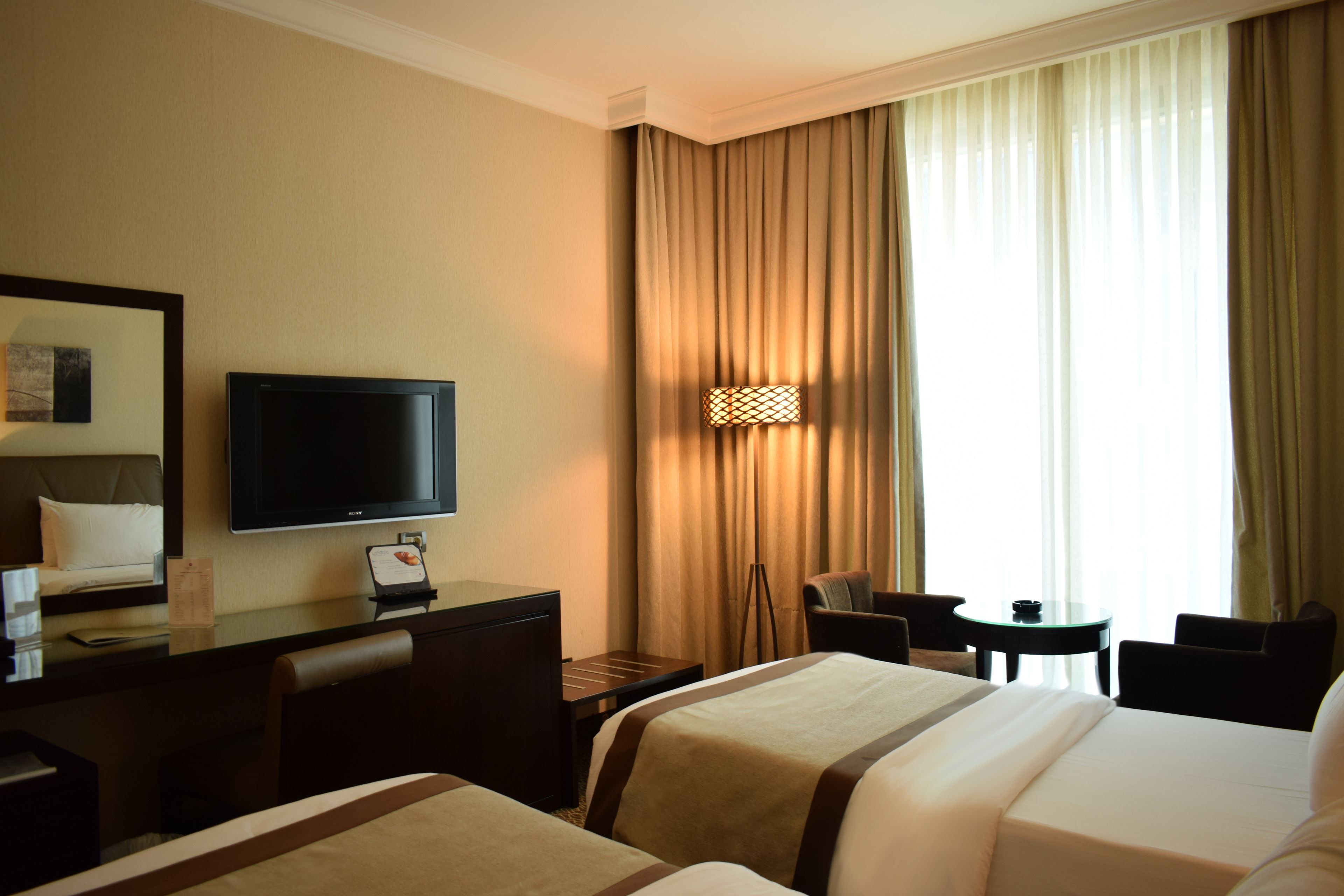 A hotel room with a bed, a desk with a television, a chair, a small table, and a window with curtains.
