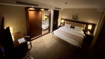 A hotel room with a bed, a desk, a chair, a wardrobe, and a nightstand with a lamp.