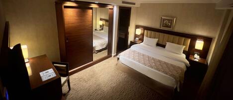 A hotel room with a bed, a desk, a chair, a wardrobe, and a nightstand with a lamp.