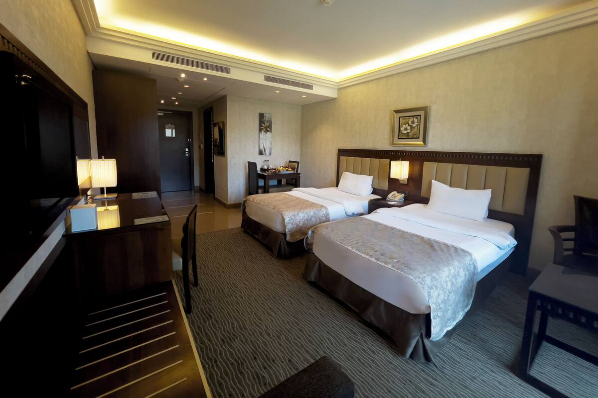 A hotel room with two beds, a desk with a lamp, a TV, and a small table.