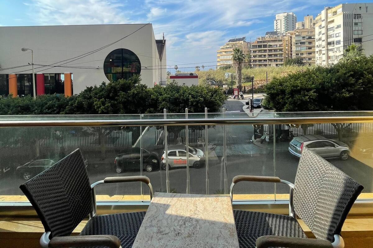 A balcony with two chairs and a table, overlooking a cityscape with buildings and a clear sky.