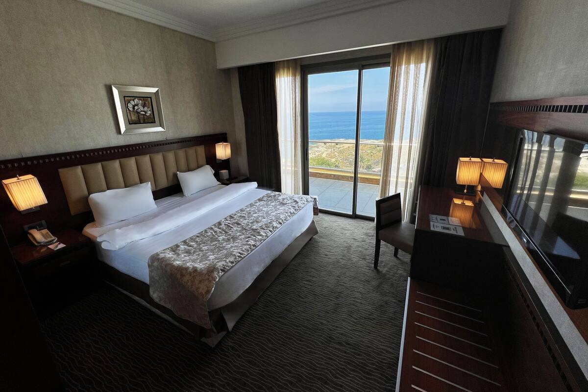 A hotel room with a large bed, a desk with a chair, a TV, a phone, and a view of the ocean through a sliding door.