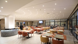 A spacious restaurant with multiple tables and chairs, a central counter, and a television screen.