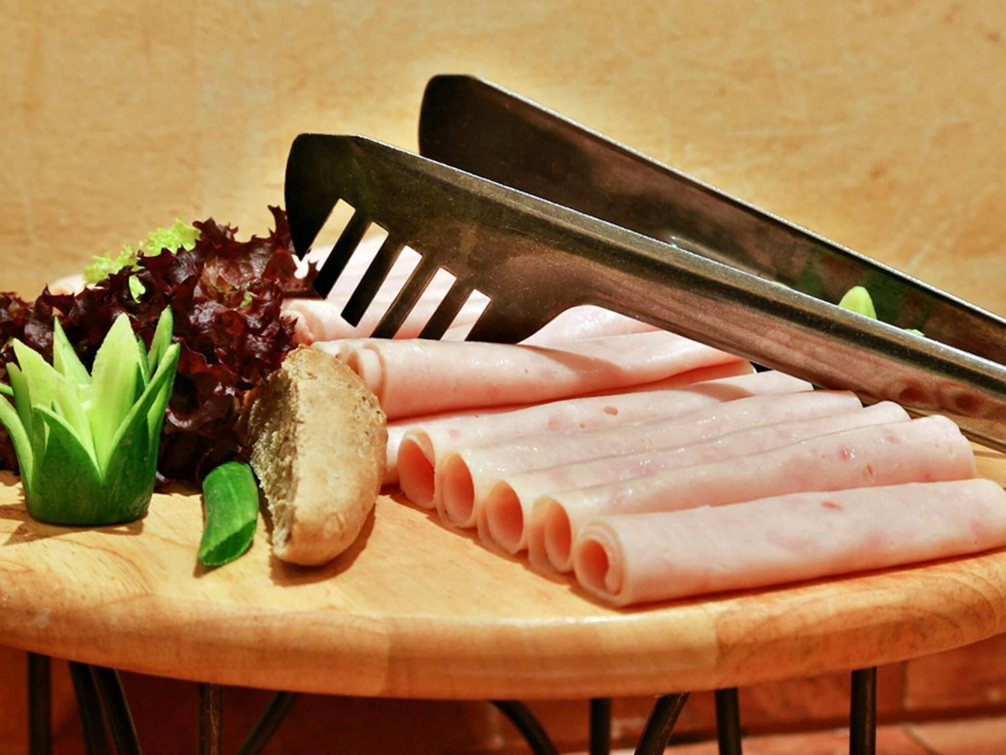 A wooden cutting board with sliced ham, a knife, a fork, and some greens.