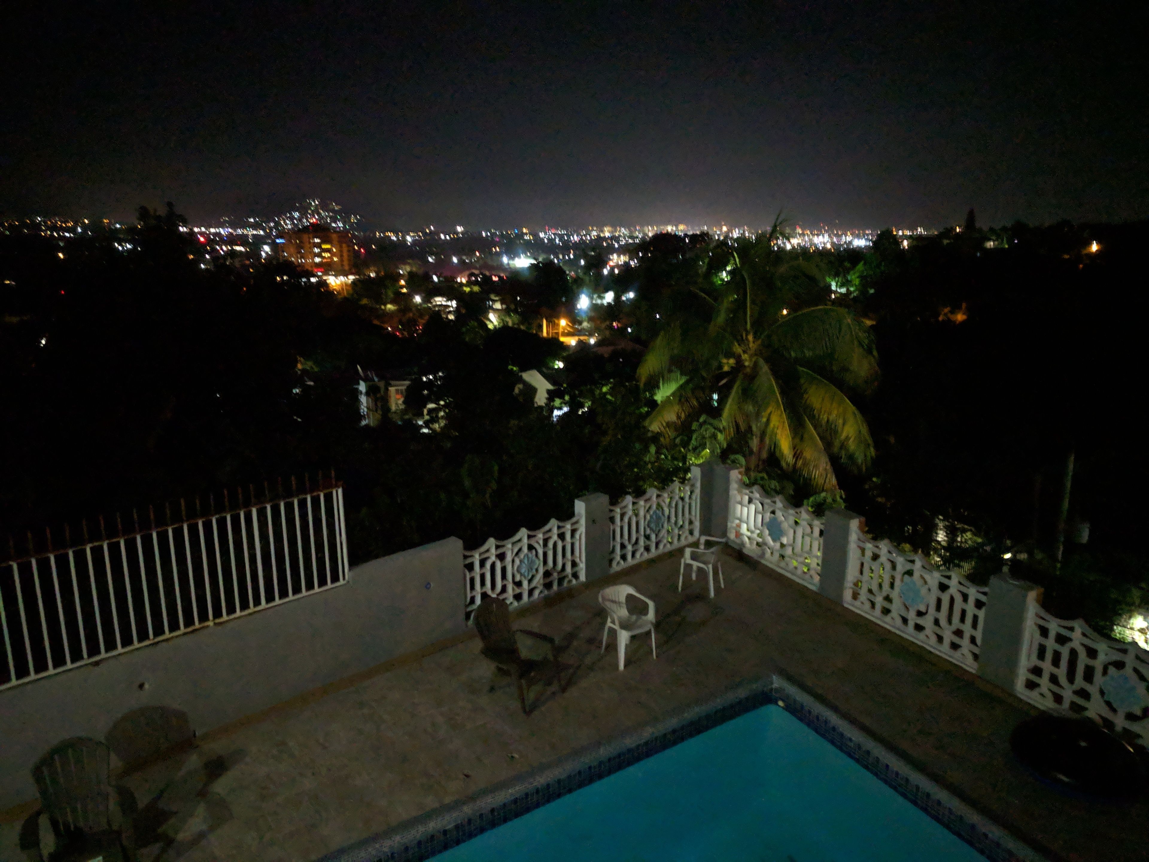 A nighttime view of a cityscape from a balcony with a swimming pool, chairs, and a palm tree.