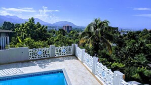 Una piscina con una valla decorativa blanca, rodeada de abundante vegetación y vistas a montañas lejanas.