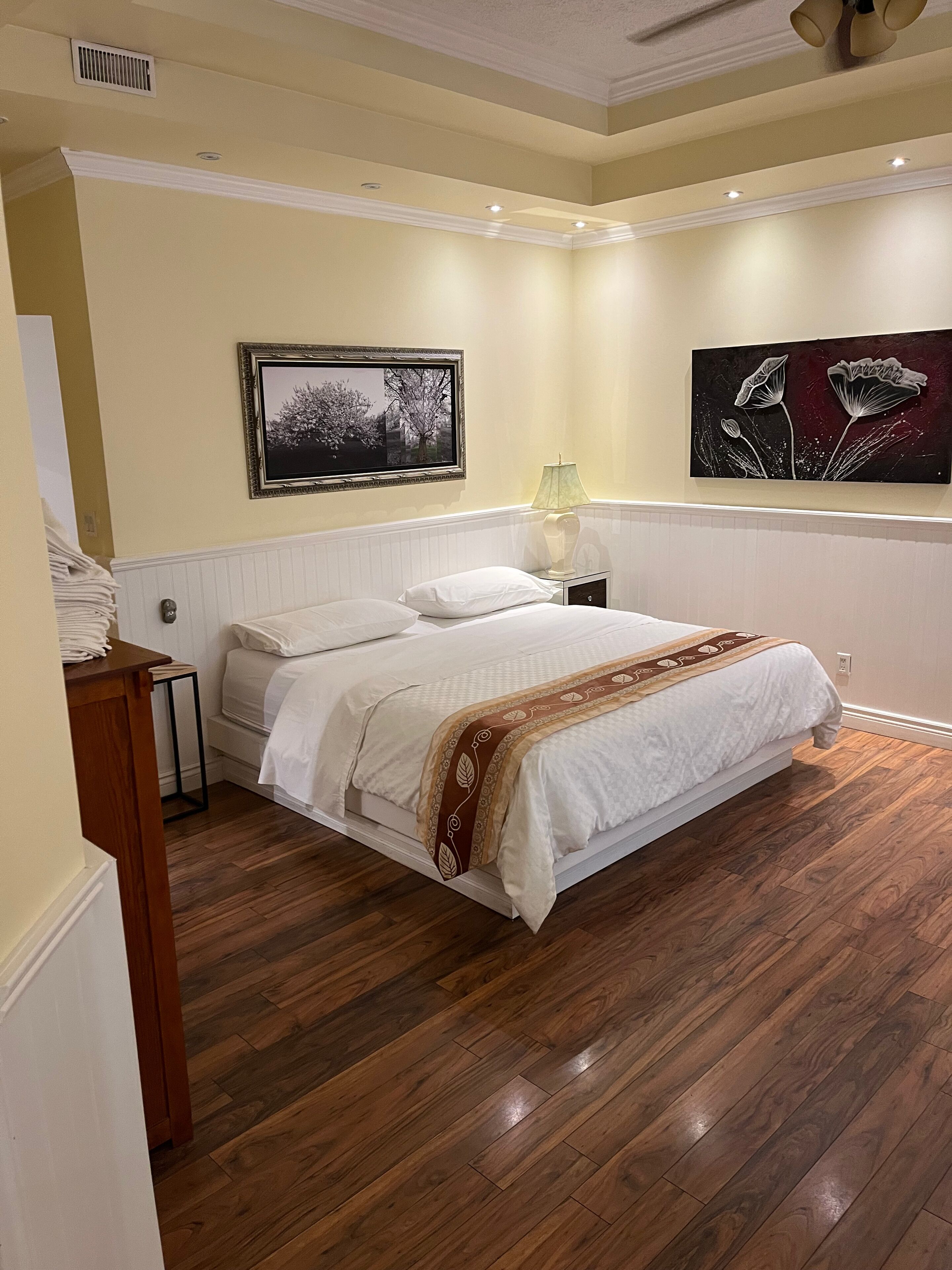 A neatly made bed in a room with wooden flooring, a white headboard, and two wall-mounted paintings.